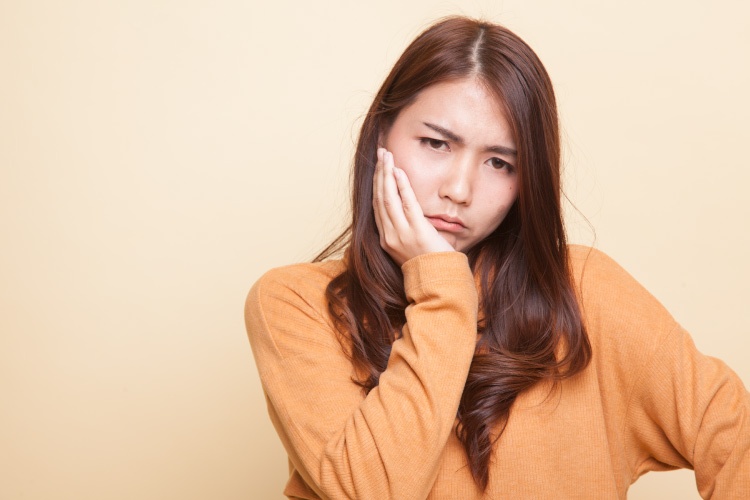 Brunette young woman wearing a gold shirt cringes and touches her cheek due to a toothache emergency