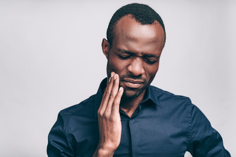 Dark-haired man wearing a navy dress shirt cringes and touches his cheek due to tooth pain