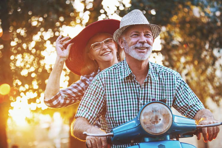 Smiling older woman & man on a scooter with the sun behind them filtered through the trees