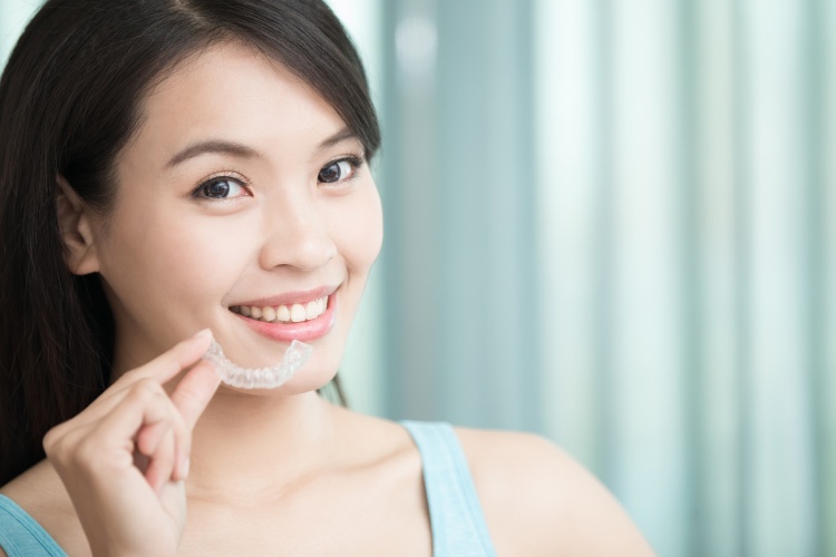 Dark haired girl smiling and holding a clear aligner like Invisalign or ClearCorrect