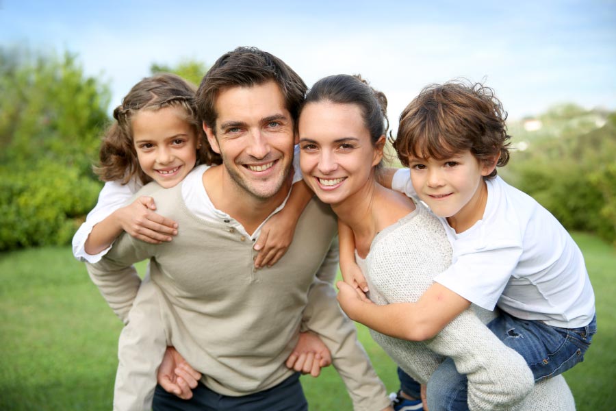 A smiling family with a mom and dad holding a young girl and young boy on their backs.